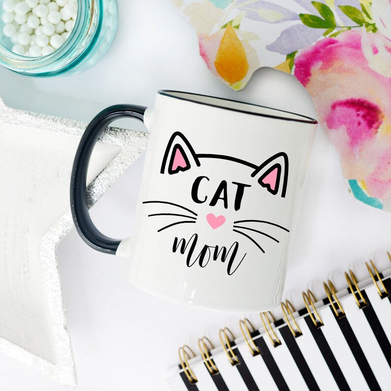 May include: White ceramic mug with a black handle and black rim. The mug has a black and white illustration of a cat face with pink ears and a pink heart nose. The text "CAT MOM" is written below the cat face.