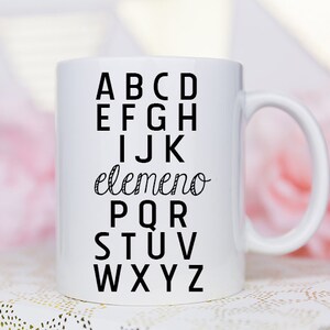 May include: White ceramic mug with a black design of the alphabet with the word "elemento" in cursive between the letters I and J.