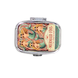 May include: A silver rectangular compact mirror with a hinged lid. The mirror's design features four mermaids with sunglasses, green tails, and blonde hair, swimming in a sea of green and pink. Text on the mirror reads "MERMAID FINS - For a Deep Sea Snack."