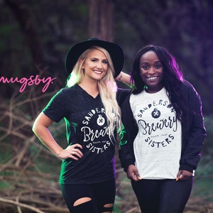 May include: Two women wearing "Sanders Brewery Sisters" t-shirts pose outdoors. One wears a black V-neck shirt and a black hat, the other a white and black raglan shirt. The logo "mugsby" is in the upper left.