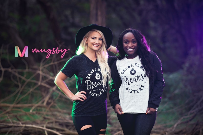 May include: Two women wearing "Sanders Brewery Sisters" t-shirts pose outdoors. One wears a black V-neck shirt and a black hat, the other a white and black raglan shirt. The logo "mugsby" is in the upper left.