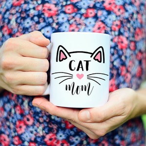 May include: White ceramic mug with a black cat face design and the text "CAT MOM" in black lettering. A pink heart is in the center of the cat's nose.