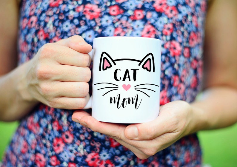 May include: White ceramic mug with a black cat face design and the text "CAT MOM" in black lettering. A pink heart is in the center of the cat's nose.