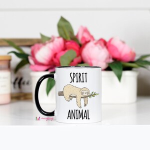 May include: White ceramic mug with a black rim and handle. The mug features a black and white illustration of a sloth hanging from a branch with the text "Spirit Animal" printed below.