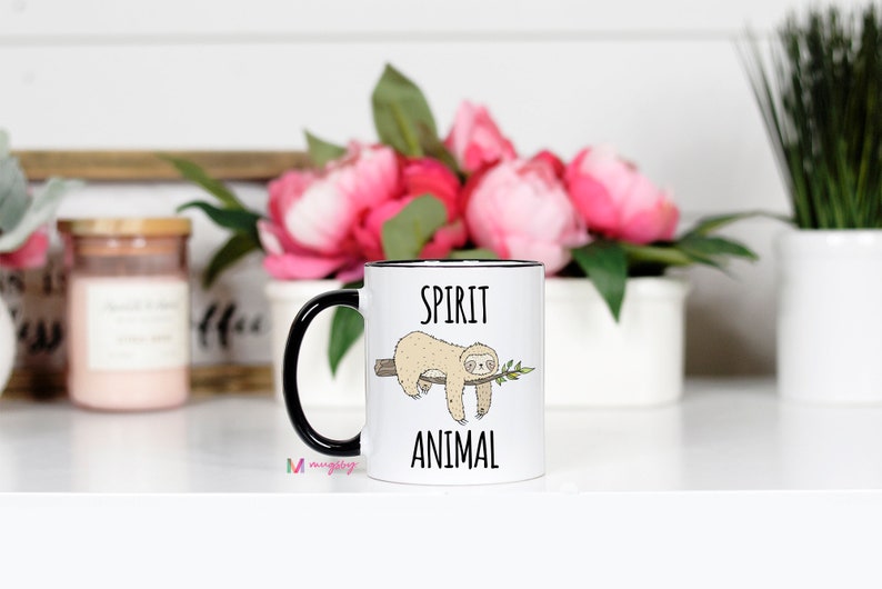 May include: White ceramic mug with a black rim and handle. The mug features a black and white illustration of a sloth hanging from a branch with the text "Spirit Animal" printed below.