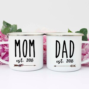 May include: Two white enamel mugs with black lettering. The mug on the left says "MOM est. 2018" with a brown arrow and floral design. The mug on the right says "DAD est. 2018" with a brown arrow.