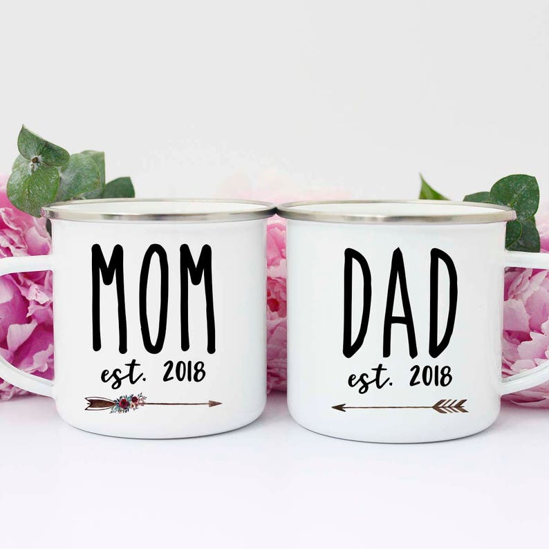 May include: Two white enamel mugs with black lettering. The mug on the left says "MOM est. 2018" with a brown arrow and floral design. The mug on the right says "DAD est. 2018" with a brown arrow.