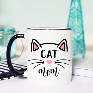 May include: White ceramic mug with a black handle. The mug has a black outline of a cat face with pink ears and a pink heart nose. The text "CAT MOM" is written below the cat face.