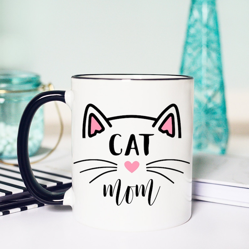 May include: White ceramic mug with a black handle. The mug has a black outline of a cat face with pink ears and a pink heart nose. The text "CAT MOM" is written below the cat face.
