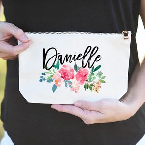 K&ouml;nnte beinhalten: Wei&szlig;e Kosmetiktasche aus Canvas mit Rei&szlig;verschluss. Die Tasche hat ein Blumenmuster mit rosa Bl&uuml;ten und gr&uuml;nen Bl&auml;ttern. Der Name "Danielle" ist in schwarzer Kursivschrift geschrieben.
