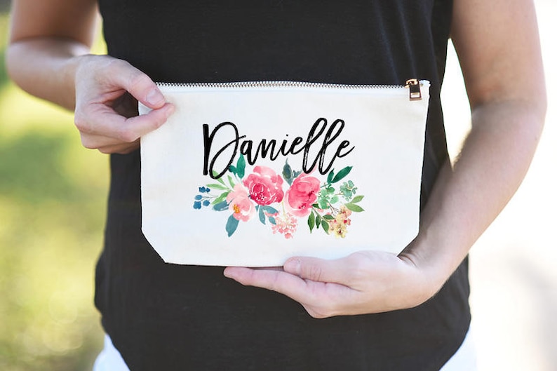 K&ouml;nnte beinhalten: Wei&szlig;e Kosmetiktasche aus Canvas mit Rei&szlig;verschluss. Die Tasche hat ein Blumenmuster mit rosa Bl&uuml;ten und gr&uuml;nen Bl&auml;ttern. Der Name "Danielle" ist in schwarzer Kursivschrift geschrieben.