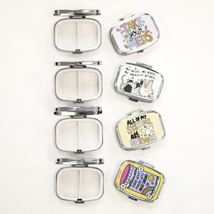 May include: A collection of rectangular, silver-colored pill cases with hinged lids. Some cases are open, revealing white interiors. Other cases feature colorful designs with text that reads "Take your meds," "My neck, my back, my anxiety attack," and "All of my systems are nervous."