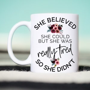 May include: White ceramic mug with the text "SHE BELIEVED SHE COULD BUT SHE WAS really tired SO SHE DIDN'T" in black script. Floral accents in burgundy and pink adorn the mug.