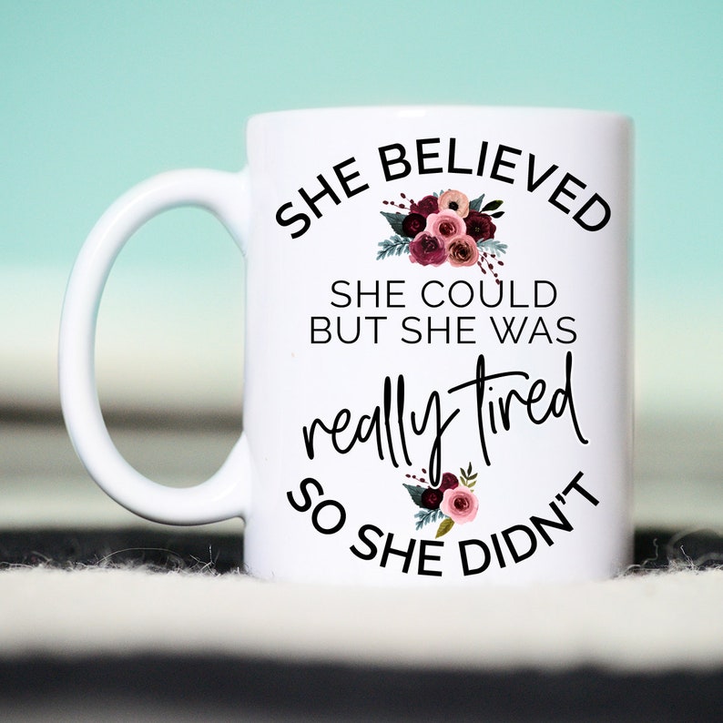 May include: White ceramic mug with the text "SHE BELIEVED SHE COULD BUT SHE WAS really tired SO SHE DIDN'T" in black script. Floral accents in burgundy and pink adorn the mug.