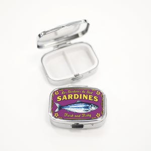 May include: A silver pill box with a purple lid featuring a sardine illustration. The lid has the words "Sardines" and "Fresh and Fishy". The pill box is open, revealing two compartments.
