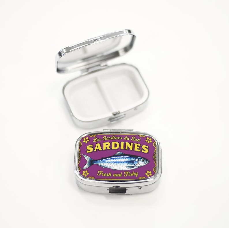 May include: A silver pill box with a purple lid featuring a sardine illustration. The lid has the words "Sardines" and "Fresh and Fishy". The pill box is open, revealing two compartments.