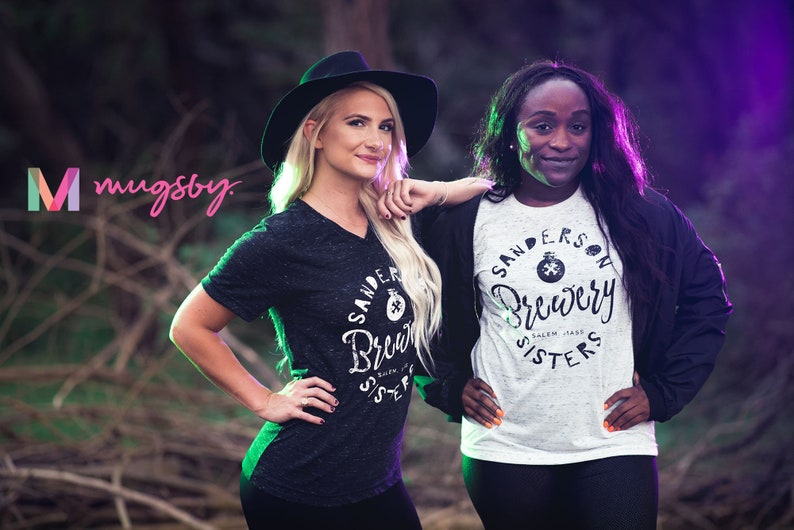 May include: Two women wearing t-shirts with the text "Sanderson Brewery Sisters" in a forest setting. One woman wears a black hat and a dark grey v-neck shirt. The other woman wears a white and grey t-shirt and a black jacket.