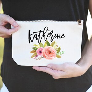 K&ouml;nnte beinhalten: Wei&szlig;e Kosmetiktasche aus Canvas mit Rei&szlig;verschluss. Die Tasche hat ein Blumenmuster mit rosa und gelben Blumen und gr&uuml;nen Bl&auml;ttern. Der Name "Katherine" ist in schwarzer Kursivschrift geschrieben.