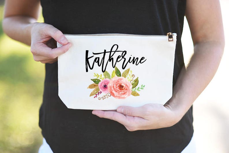 K&ouml;nnte beinhalten: Wei&szlig;e Kosmetiktasche aus Canvas mit Rei&szlig;verschluss. Die Tasche hat ein Blumenmuster mit rosa und gelben Blumen und gr&uuml;nen Bl&auml;ttern. Der Name "Katherine" ist in schwarzer Kursivschrift geschrieben.