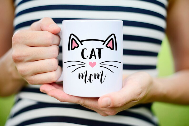May include: White ceramic mug with a black cat face design and the text "CAT MOM" in black lettering. A pink heart is in the center of the cat's face.