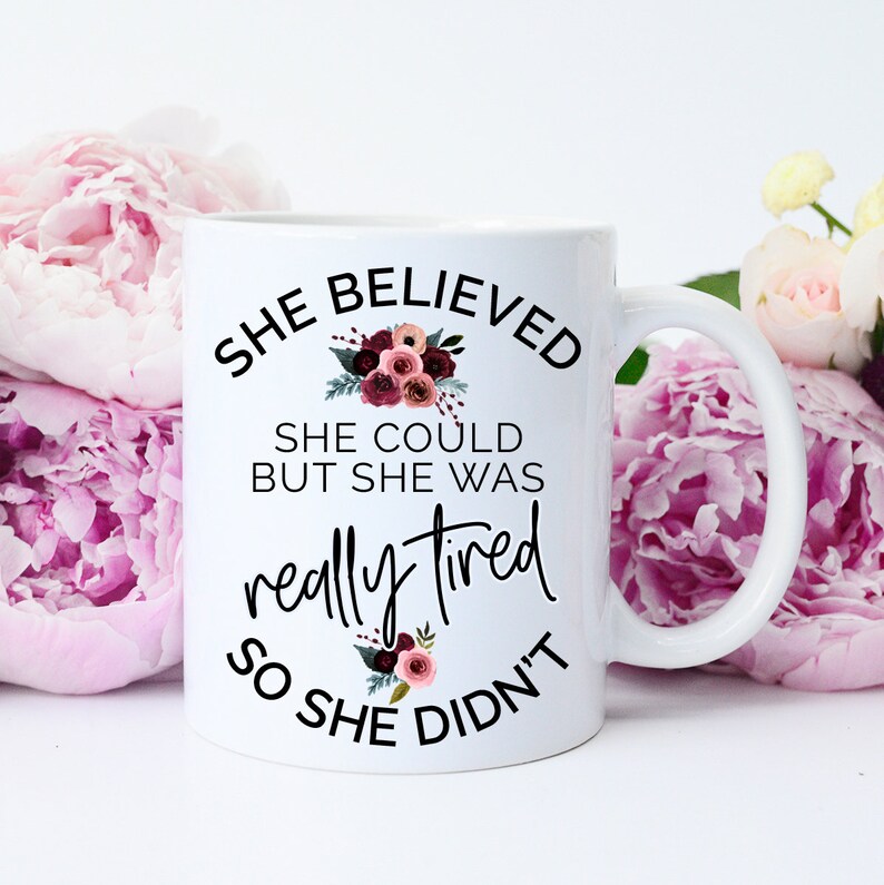 May include: White ceramic mug with the text "SHE BELIEVED SHE COULD BUT SHE WAS really tired SO SHE DIDN'T" in black script. Floral accents in burgundy and pink surround the text. The mug has a curved handle and is surrounded by pink flowers.