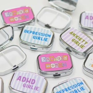 May include: A collection of small, rectangular, silver-colored pill boxes. Each box has a colorful, patterned lid with text such as "Enraged Woman," "Depression Girlie," and "ADHD Girlie." The boxes are open and closed, revealing white interiors.