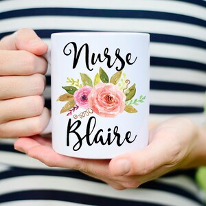 Puede incluir: Taza de caf&eacute; de cer&aacute;mica blanca con un dise&ntilde;o floral y el texto "Nurse Blair" en letras negras.