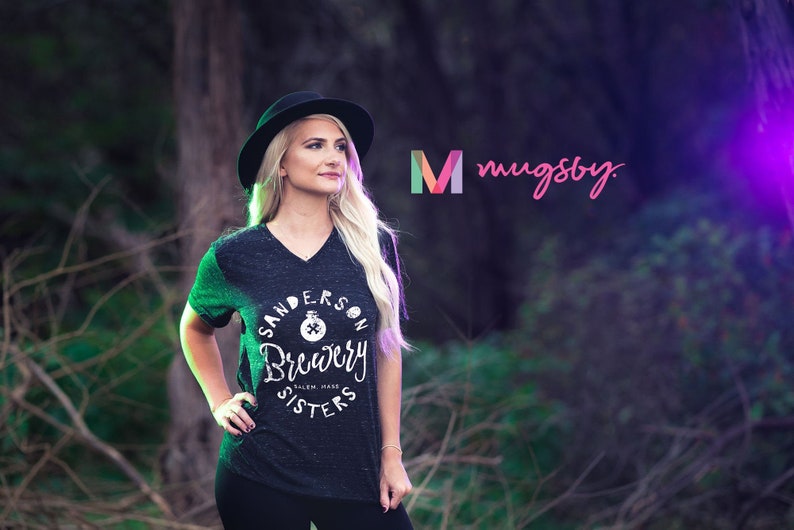 May include: Dark gray v-neck t-shirt with the text "Sanderson Brewery Sisters" in a circular design. The shirt is worn by a person wearing a black hat. The image also includes the logo "mugsby."