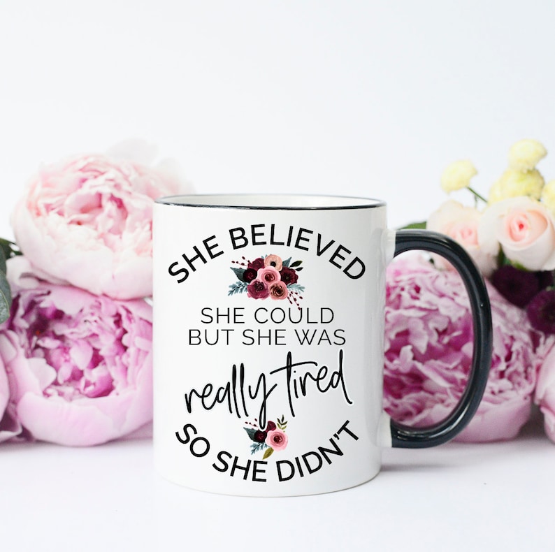 May include: White ceramic mug with a black handle and rim, featuring the text "She Believed She Could But She Was Really Tired So She Didn't." The design includes floral accents in shades of burgundy and green.