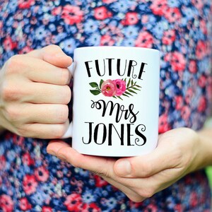 May include: White ceramic mug with black text that reads "Future Mrs. Jones" with a floral design featuring pink flowers and green leaves.