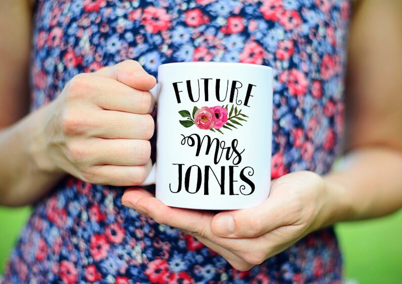 May include: White ceramic mug with black text that reads "Future Mrs. Jones" with a floral design featuring pink flowers and green leaves.