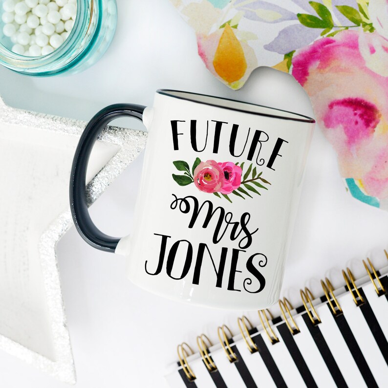 May include: White ceramic mug with a black handle and black rim. The mug has a floral design with pink flowers and green leaves. The text "FUTURE Mrs JONES" is printed in black on the mug.