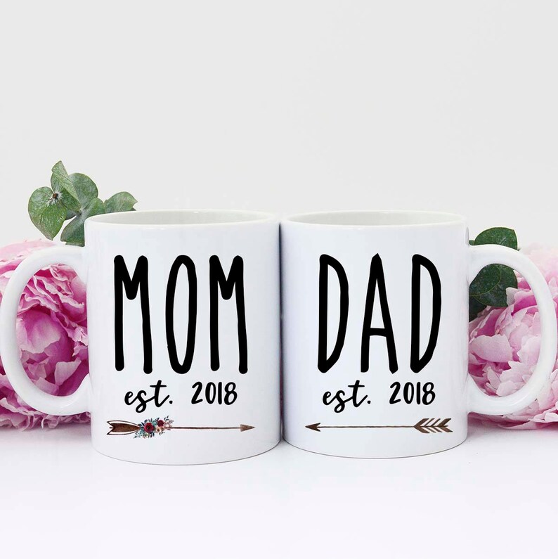 May include: Two white ceramic mugs with black text. The mug on the left says "MOM est. 2018" with a small arrow and floral design. The mug on the right says "DAD est. 2018" with a small arrow.
