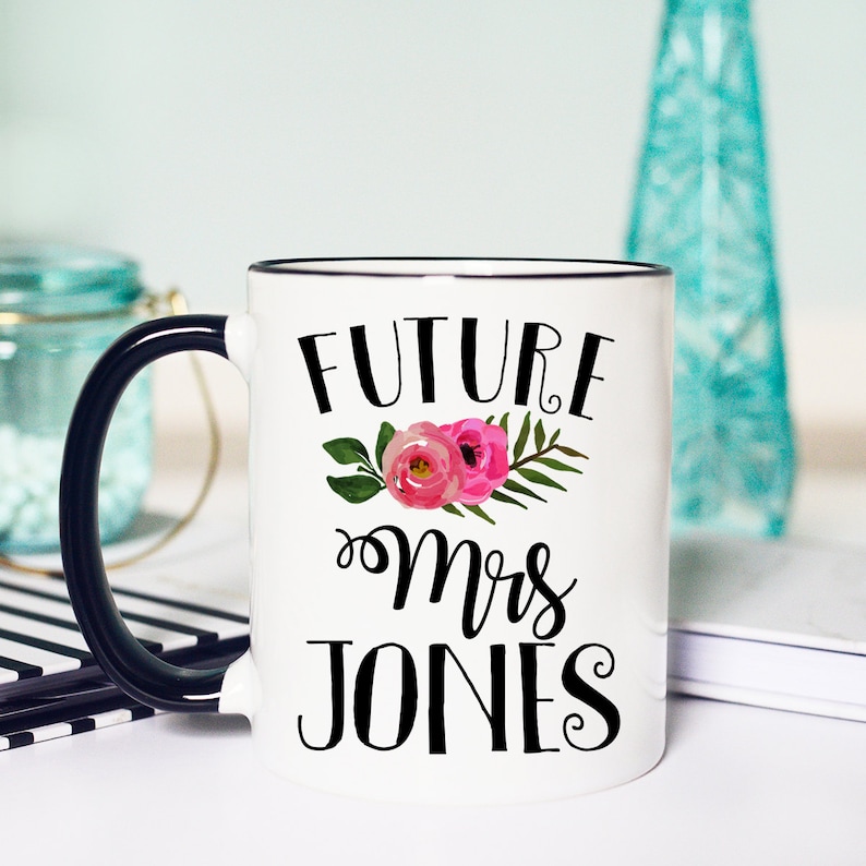 May include: White ceramic mug with a black rim and handle. The mug has black text that reads "FUTURE Mrs JONES" with a floral design in pink and green.