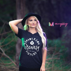 May include: Dark gray v-neck t-shirt with the text "Sanderson Brewery Sisters" in white. The shirt features a round graphic with a cauldron and the text "Salem, Mass". A woman wearing a black hat is in the image.
