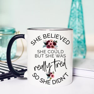 May include: White ceramic mug with a black handle and rim. The mug features the text "She Believed She Could But She Was Really Tired So She Didn't" in black script, with floral accents in burgundy and pink. A humorous gift.