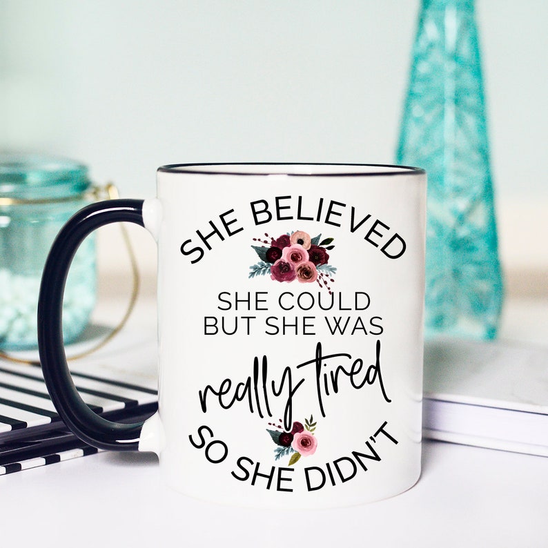 May include: White ceramic mug with a black handle and rim. The mug features the text "She Believed She Could But She Was Really Tired So She Didn't" in black script, with floral accents in burgundy and pink. A humorous gift.