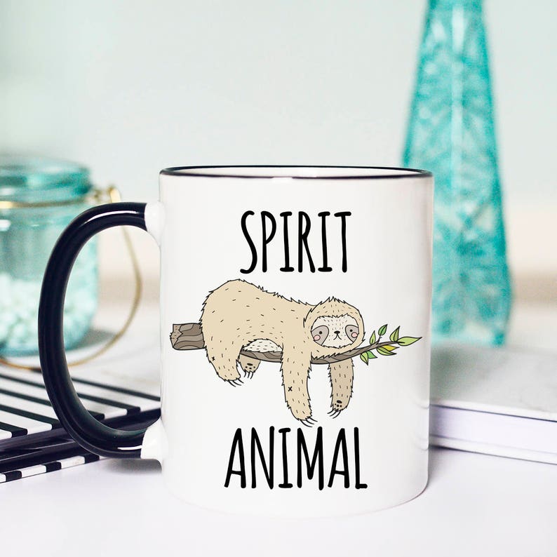 May include: White ceramic mug with a black handle. The mug has a black rim and features a cartoon sloth hanging from a branch with the text "SPIRIT ANIMAL" printed on the mug.