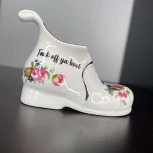 May include: White ceramic boot-shaped vase with a curved handle. The vase features floral patterns in pink, yellow, and blue. The text "Fuck off ya boot" is written on the side in black.