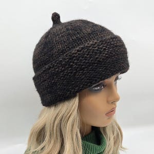 May include: A dark brown knit beanie with a small, rounded top detail. The hat has a folded brim and a textured appearance. The beanie is shown on a mannequin with blonde hair and a green sweater.