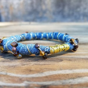 May include: A handmade bracelet featuring a denim fabric base, wrapped with gold-colored wire. The bracelet is adorned with brown and amber-colored beads. The bracelet is a unique piece of jewelry.