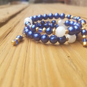 May include: A blue and gold beaded wrap bracelet with white beads. The bracelet is coiled on a wooden surface.