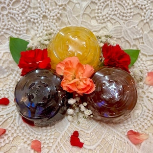 May include: Three round soaps, one yellow, two brown, arranged on a white lace background with red and pink flowers.