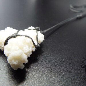 May include: A white coral pendant tied with black cord. The coral is a cluster of small, white, textured pieces. The cord is tied in a knot at the top of the pendant.