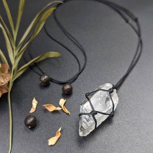 May include: A clear quartz crystal pendant wrapped in black cord and hanging from a black cord necklace. The necklace is on a grey surface with brown seeds and dried leaves.