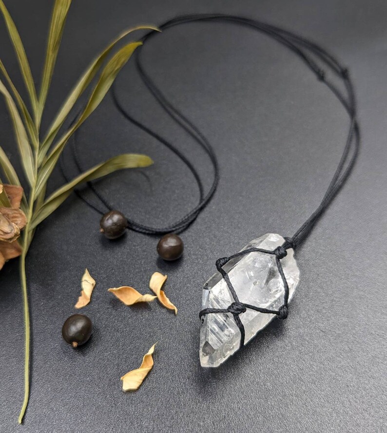 May include: A clear quartz crystal pendant wrapped in black cord and hanging from a black cord necklace. The necklace is on a grey surface with brown seeds and dried leaves.