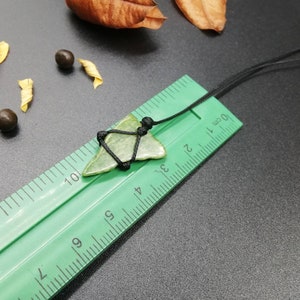 Pu&ograve; includere: Un ciondolo a forma di punta di freccia in pietra verde su un cordoncino nero, misurato su un righello verde con segni in centimetri.