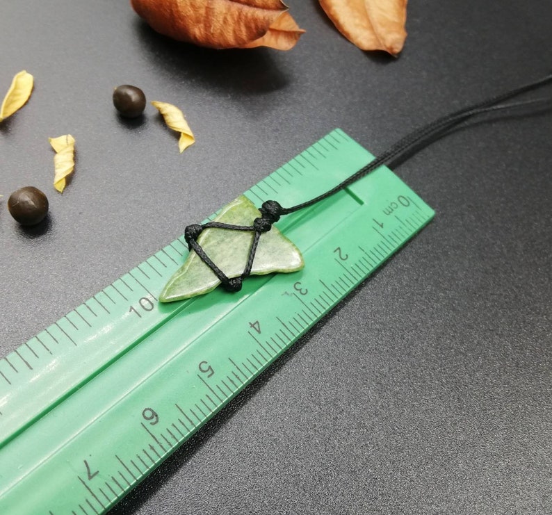 Pu&ograve; includere: Un ciondolo a forma di punta di freccia in pietra verde su un cordoncino nero, misurato su un righello verde con segni in centimetri.