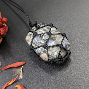 May include: A black cord necklace with a grey and blue stone pendant wrapped in a black macrame knot.