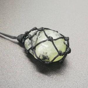 May include: A green and black crystal stone pendant is wrapped in a black macrame cord. The pendant is tied to a black cord.
